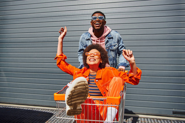 Χαρούμενοι νέοι αφροαμερικανοί καλύτεροι φίλοι με γυαλιά ηλίου και λαμπερά ρούχα διασκεδάζουν με το καλάθι αγορών και περνούν χρόνο κοντά στο κτίριο στο παρασκήνιο, φίλοι που κάνουν παρέα - Φωτογραφία, εικόνα