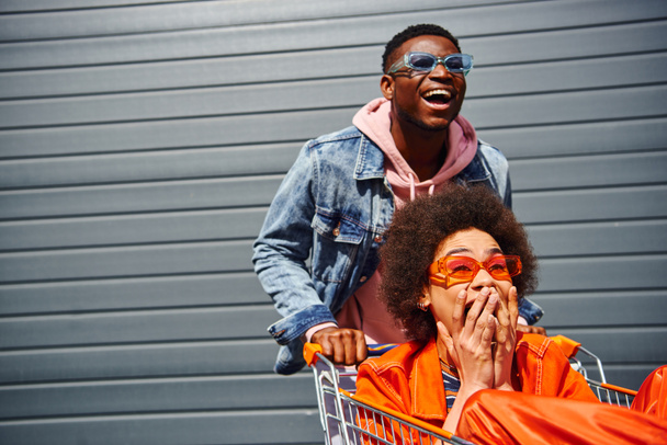 Cheerful young african american man in sunglasses having fun with scared best friend in trendy outfit and shopping cart near building on urban street, friends hanging out together - Фото, изображение