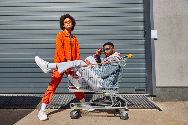 Full length of trendy and confident young african american best friends in sunglasses and bright outfits looking at camera and posing with shopping cart on urban street, friends with stylish vibe - Foto, imagen