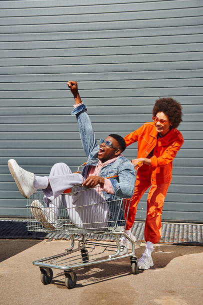 Full length of smiling young african american woman in sunglasses and bright outfit having fun with excited best friend sitting in shopping cart near building on urban street, stylish friends in city - Photo, Image