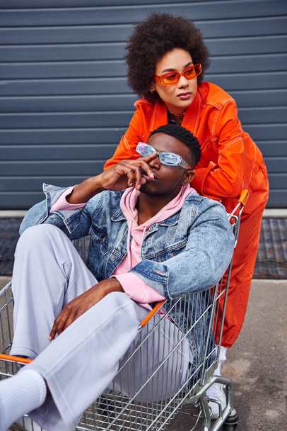 Modern young african american woman in sunglasses and bright outfit looking away while standing near best friend sitting in shopping cart and building on urban street, stylish friends in city - Fotografie, Obrázek