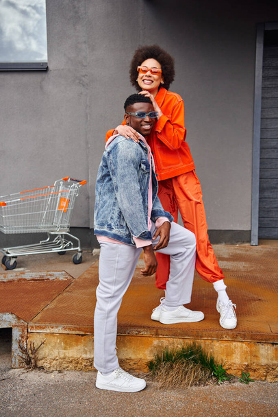 Full length of cheerful young african american woman in bright outfit and sunglasses hugging best friend in denim jacket while standing on stairs near building outdoors, stylish friends in city - Zdjęcie, obraz