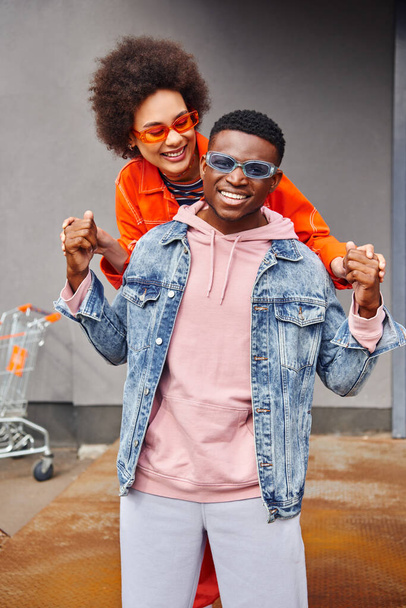 Happy young african american woman in sunglasses holding hands of trendy best friend in denim jacket while standing together near building on urban street, stylish friends in city concept - Foto, imagen