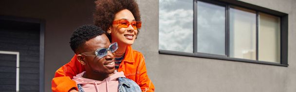 Positiva giovane donna afroamericana in occhiali da sole e vestito moderno che abbraccia il migliore amico e distoglie lo sguardo mentre si trova insieme vicino all'edificio sulla strada urbana, amici alla moda in città, banner  - Foto, immagini