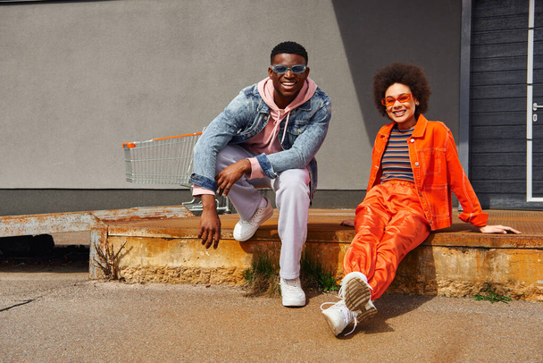 Full length of cheerful and trendy young african american best friends in sunglasses and modern outfits looking at camera while posing on rusty stairs near building outdoors, stylish friends in city - Foto, Bild