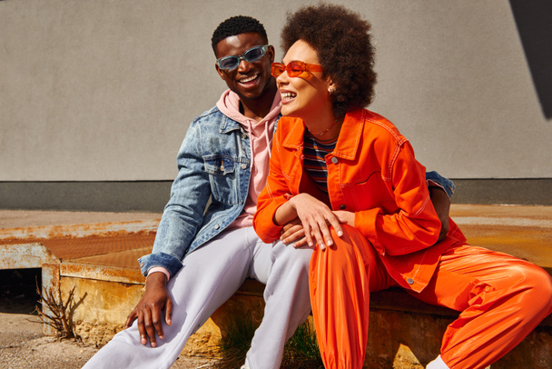 Fröhliche junge afrikanisch-amerikanische beste Freunde in trendigen Outfits und Sonnenbrille, die lachen und Spaß haben, während sie auf rostigen Treppen in der Nähe von Gebäuden an der Stadtstraße sitzen, trendige Freunde in urbanen Umgebungen - Foto, Bild