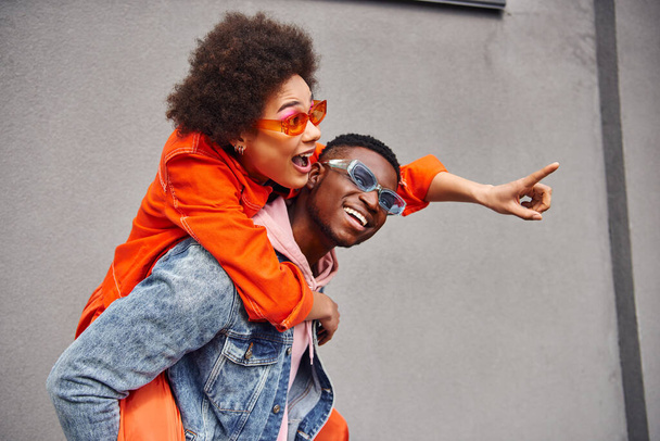 Ενθουσιασμένη νεαρή Αφροαμερικανή γυναίκα με γυαλιά ηλίου και κομψό ντύσιμο που δείχνει με το δάχτυλο, ενώ piggybacking σε φίλο και στέκεται κοντά σε κτίριο σε αστικό δρόμο, trendy φίλους σε αστικό περιβάλλον - Φωτογραφία, εικόνα