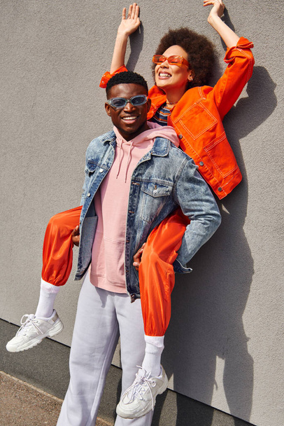 Positive and trendy young african american man in denim jacket holding best friend in sunglasses and bright outfit and standing near building on urban street, trendy friends in urban settings - Photo, Image
