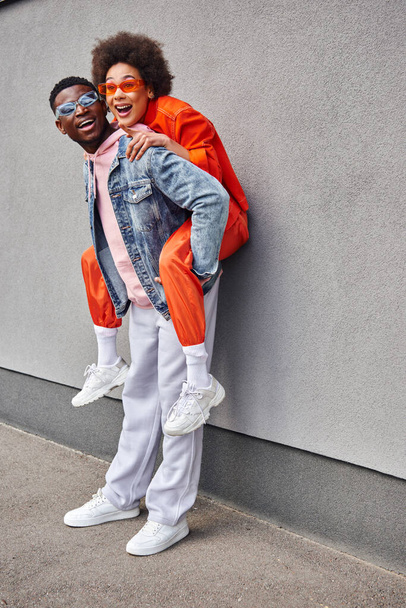 Excited african american woman in sunglasses and stylish outfit pointing with finger and piggybacking on best friend while standing near building on urban street, friends with trendy aesthetic - 写真・画像