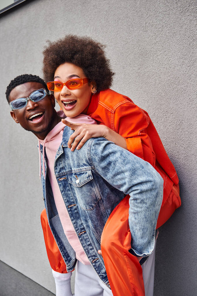 Cheerful young african american woman in stylish sunglasses pointing with finger while piggybacking on best friend and standing near building on urban street, friends with trendy aesthetic - Photo, Image