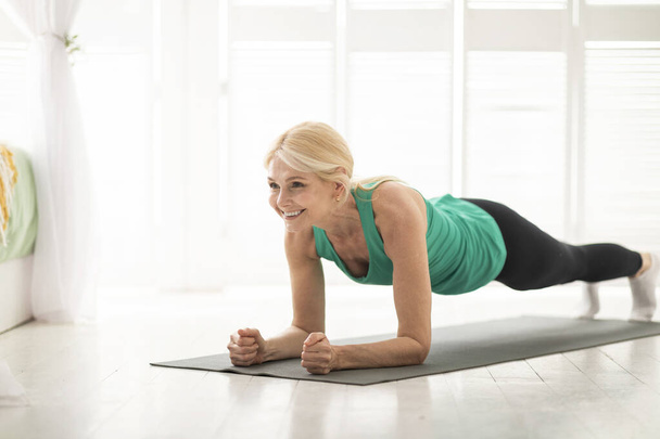 Home training concept. Smiling senior woman doing elbow plank on yoga mat in living room, sporty mature lady doing fitness workout routine, exercising indoors, strengthening her core muscles - Zdjęcie, obraz