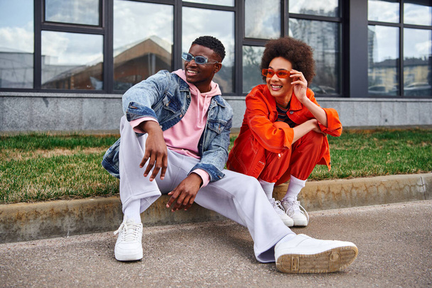 Tam boy gülümseyen ve modaya uygun Afro-Amerikalı güneş gözlüklü en iyi arkadaşlar bulanık şehir caddesinde yan yana otururken yan yana oturuyorlar, modaya uygun estetik arkadaşları - Fotoğraf, Görsel