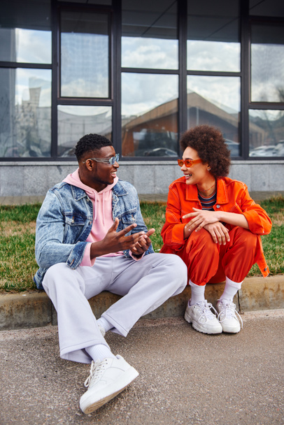 Vidám fiatal afro-amerikai legjobb barátok napszemüvegben és fényes ruhák beszélgetnek, miközben időt és ül a határon a városi utcán a háttérben, barátok trendi esztétikai - Fotó, kép