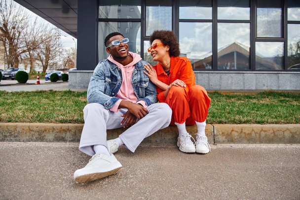 Pozytywna młoda Afroamerykanka w okularach przeciwsłonecznych i jasnym stroju rozmawia z najlepszym przyjacielem spędzając razem czas i siedząc na granicy na ulicy miejskiej, znajomi z modną estetyką - Zdjęcie, obraz