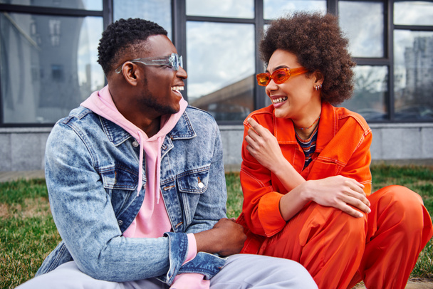 Neşeli, Afro-Amerikalı, güneş gözlüklü ve şık kıyafetler giyen en iyi arkadaşlar, şehir caddesinde bulanık binanın yanında otururken sohbet ediyorlar. - Fotoğraf, Görsel