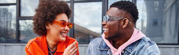 Mosolygó fiatal afro-amerikai nő napszemüvegben és fényes ruhát beszél a legjobb barátja farmerdzsekiben, miközben időt tölt a városi utcán, barátok trendi esztétikai, banner  - Fotó, kép