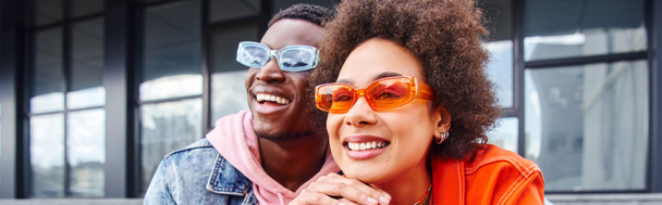 Mulher afro-americana jovem positiva com cabelo natural vestindo óculos de sol elegantes e olhando para longe perto melhor amigo enquanto passar o tempo na rua urbana, amigos com estética na moda, banner  - Foto, Imagem