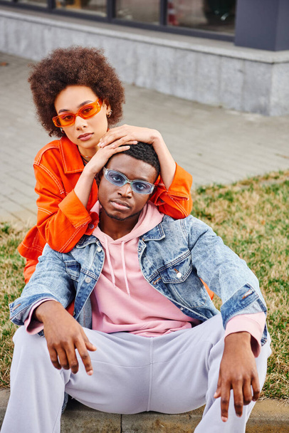 Módní mladá africká americká žena v slunečních brýlích a světlé oblečení objímání a pózování s nejlepším přítelem v džínové bundě a při pohledu na kameru na městské ulici, stylové přátelé těší společnost - Fotografie, Obrázek
