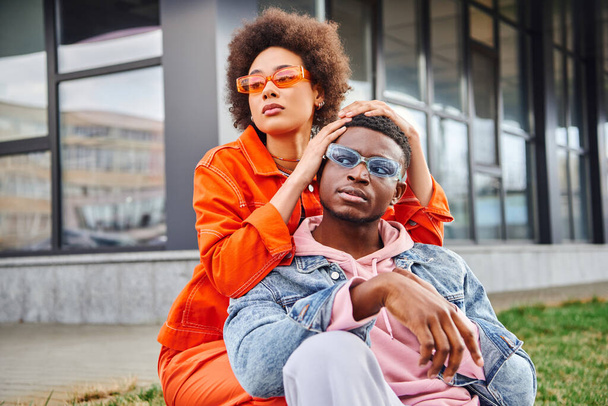 Confident and stylish young african american woman in sunglasses and bright outfit posing with best fried and looking away while spending time on urban street, stylish friends enjoying company - Foto, afbeelding