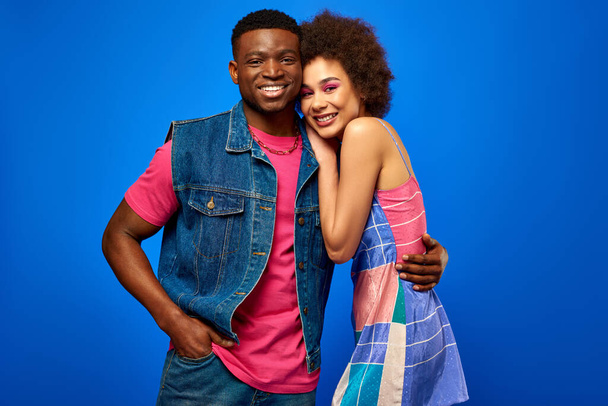 Vrolijke en stijlvolle jonge Afrikaanse Amerikaanse man in denim vest knuffelen beste vriend in sundress en kijken naar de camera terwijl geïsoleerd op blauwe, stijlvolle vrienden stralend vertrouwen - Foto, afbeelding