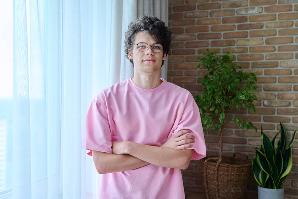 Портрет красивого парня 18-19 лет со скрещенными руками в интерьере дома. Кучерявый молодой студент колледжа в розовой футболке с очками, смотрящий в камеру. Молодежь, возраст, образ жизни - Фото, изображение