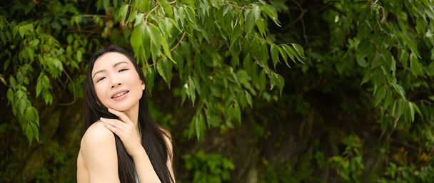 Κοντινό πλάνο υπαίθρια πορτρέτο Όμορφη νεαρή κινεζική ασιατική γυναίκα τέλειο δέρμα ποζάροντας έξω ηλιόλουστη μέρα του καλοκαιριού πράσινο φύλλωμα. Φυσική περιποίηση προσώπου. Καλλυντικά, περιποίηση δέρματος και σπα. Έννοια αραίωσης - Φωτογραφία, εικόνα