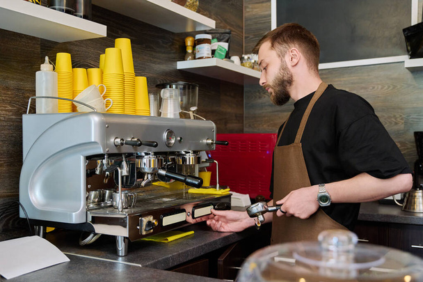 Male barista preparing coffee in coffee shop, bearded young man worker in an apron standing near coffee machine. Small business, food service occupation, coffee shop, staff, work concept - Foto, afbeelding