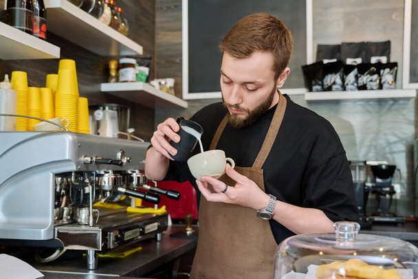 Male barista preparing coffee in coffee shop, bearded young man worker in an apron standing near coffee machine. Small business, food service occupation, coffee shop, staff, work concept - Photo, Image