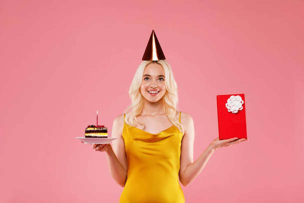 Счастливая молодая женщина в шляпе на день рождения, с тортом и красной подарочной коробкой, позирует изолированно на розовом фоне, студия снимает. Концепция праздничного праздника - Фото, изображение