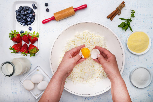 cook adds eggs to the cottage cheese to bake a casserole. Flour, eggs, rolling pin, butter, milk and strawberries on a white table. View from above. - Photo, image