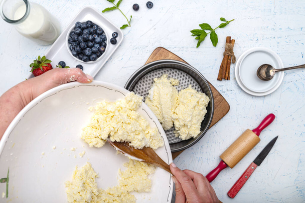The chef puts the cottage cheese in a casserole baking dish. Flour, eggs, rolling pin, butter, milk and strawberries on a white table. View from above. - Фото, зображення