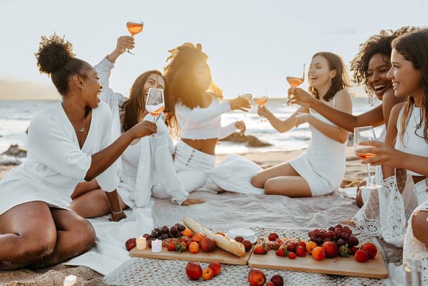 Grupo de mulheres felizes amigas do sexo feminino fazendo um piquenique aconchegante de verão com vinho ou champanhe, tendo festa de galinha na costa, copos e bate-papo - Foto, Imagem