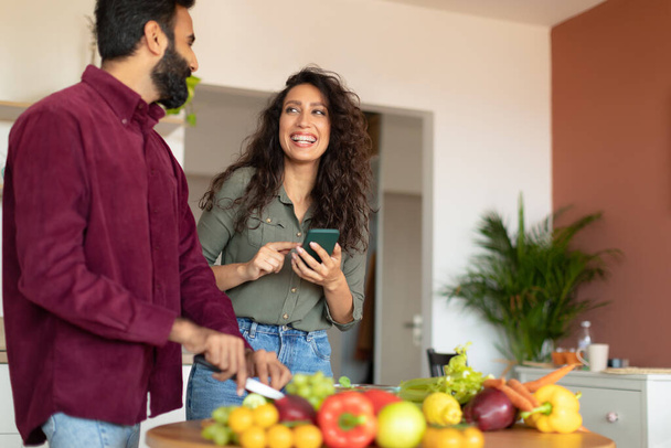 Happy arab couple cooking together, woman reading recipe on smartphone while man cutting vegetables for salad at kitchen interior, copy space. Excited spouses making lunch - Foto, Imagen