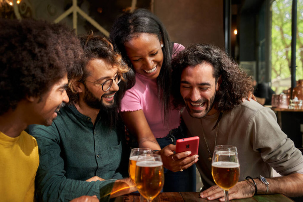 Mladá multietnická skupina lidí se dívá na mobil a usmívá se v baru. Dívka ukazuje svůj telefon svým přátelům. Shromáždění si společně užívá sociálních médií. Millennials připojen k smartphonu - Fotografie, Obrázek