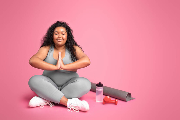 Mujer negra de gran tamaño en ropa deportiva practicando yoga, meditación, disfrutando de la pérdida de peso, sentado en el fondo del estudio rosa, espacio libre. Vida sana, cuidado del cuerpo y de la salud, entrenamiento muscular - Foto, imagen