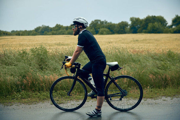 Young bearded man, cyclist in uniform, helmet and glasses standing with bicycle on road around field background. Concept of sport, hobby, leisure activity, training, health, speed, endurance, ad - Foto, imagen