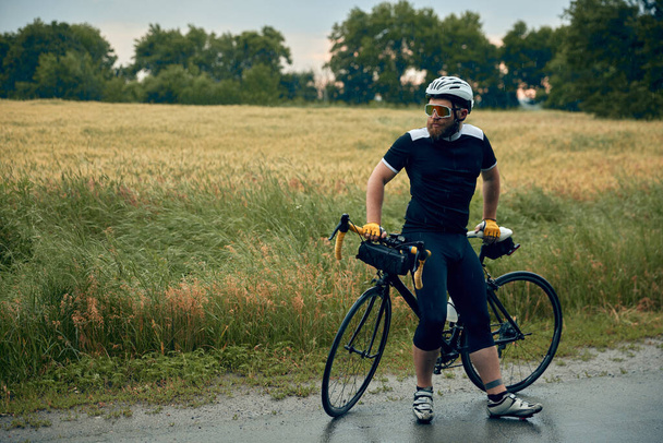 Young bearded man, cyclist in uniform, helmet and glasses sitting on bicycle on road around field background. Concept of sport, hobby, leisure activity, training, health, speed, endurance, ad - Φωτογραφία, εικόνα