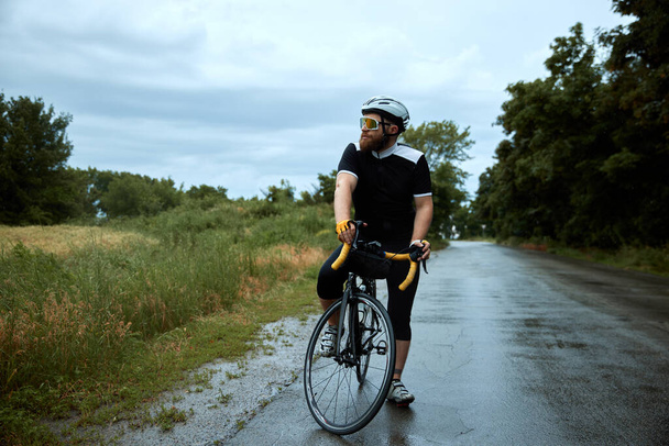 Sportive, young, bearded man, cyclist in uniform, helmet and glasses posing on bike, riding on road in chill evening. Concept of sport, hobby, leisure activity, training, health, speed, endurance, ad - Foto, immagini
