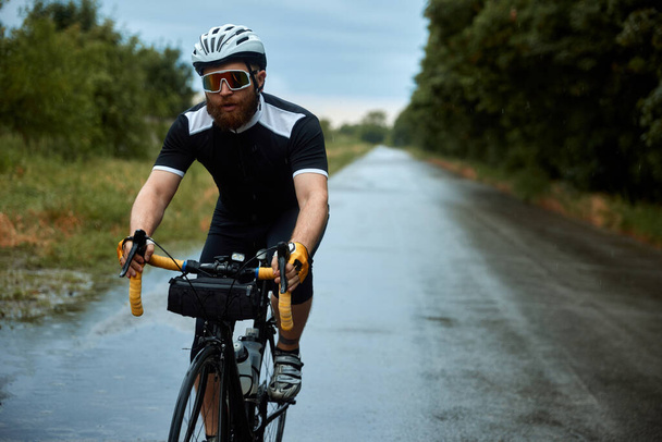 Concentrated, young, bearded man, cyclist in uniform, helmet and glasses riding bike on road in chill evening. Concept of sport, hobby, leisure activity, training, health, speed, endurance, ad - Fotografie, Obrázek