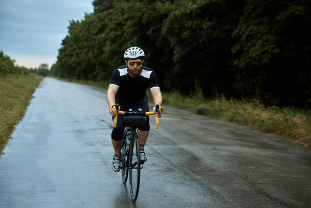 Sportive, young, bearded man, cyclist in uniform, helmet and glasses riding bike on wet road in chill evening after rain. Sport, hobby, leisure activity, training, health, speed, endurance, ad concept - Photo, Image