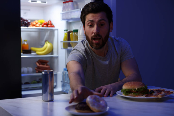 Man taking donut in kitchen at night. Bad habit - Фото, изображение