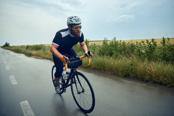 Bearded young man, cyclist in helmet, glasses and uniform riding bike on wet road in cloudy chill evening. Concept of sport, hobby, leisure activity, training, health, speed, endurance, ad - Photo, Image