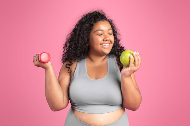 Concept d'alimentation saine. Heureuse femme positive corps noir tenant haltère et pomme dans les mains, profitant de fruits frais et d'entraînement, debout sur fond de studio rose - Photo, image