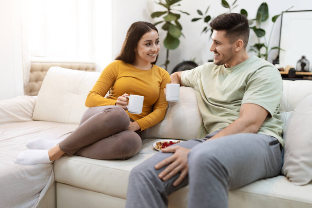 Happy attractive millennial couple drinking coffee at home. Cheeful young man and woman wearing comfy homewear sitting on couch in living-room, drinking tea and have conversation, copy space - Photo, image