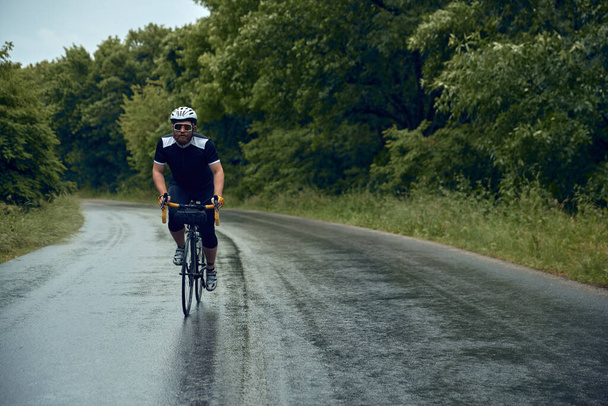 Sportive, young, bearded man, cyclist in helmet, glasses and uniform riding bike on wet road in cloudy chill evening. Concept of sport, hobby, leisure activity, training, health, speed, endurance, ad - Photo, Image