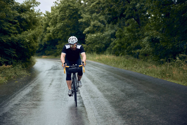Sportive, young, bearded man, cyclist in helmet, glasses and uniform riding bike on wet road in cloudy chill evening. Concept of sport, hobby, leisure activity, training, health, speed, endurance, ad - Foto, Imagem