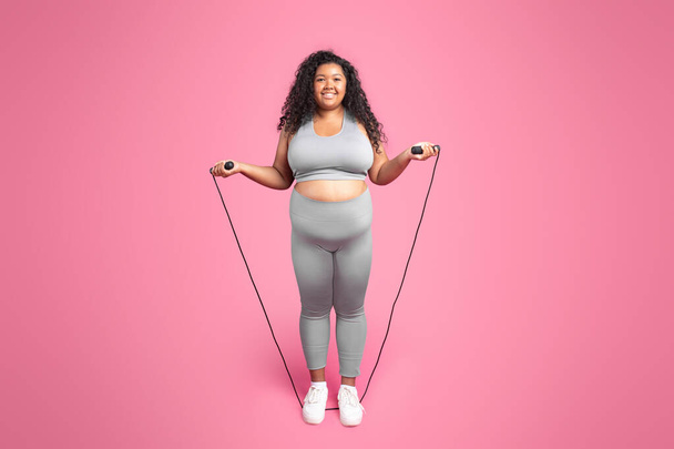 Positive black body positive woman in sportswear posing with skipping rope, doing fitness training, standing over pink background, full length, copy space - 写真・画像