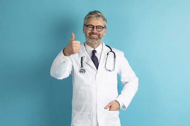 カメラで親指を表示し、笑顔、青い背景に隔離された医療用作業服の医者で良い陽気なハンサムな白髪の中年の男性。健康診断医療保険 - 写真・画像