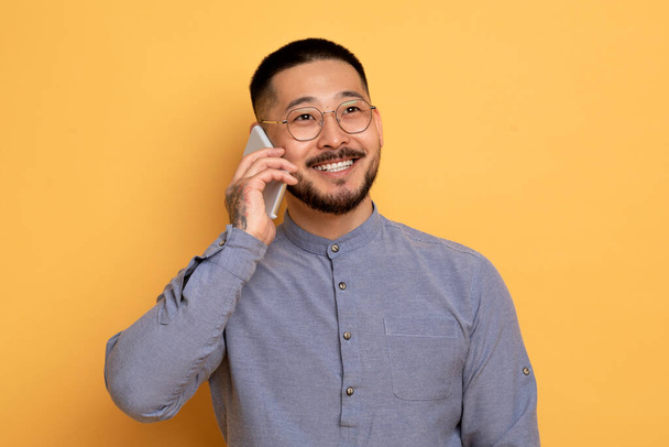 Puhelu. Onnellinen nuori aasialainen mies puhuu matkapuhelimella, iloinen tuhatvuotinen kaveri, jolla on miellyttävä puhelinkeskustelu, nauttii nykyaikaisesta viestinnästä, seisoo keltaisen studion taustalla, kopioi tilaa - Valokuva, kuva