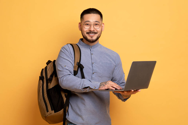 Portrait Of Young Asian Male Student With Backpack And Laptop Standing Over Yellow Studio Background, Happy Millennial Man Holding Computer And Smiling At Camera, Enjoying Online Education - Φωτογραφία, εικόνα
